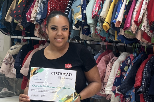 Chanelle is pictured in Ealing Baby Bank holding the certificate and glass trophy that she was awarded. Behind her is a display of colourful clothes for babies and young children.
