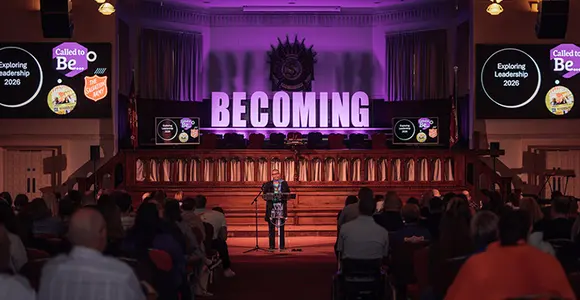 Speaker standing at a podium in front of an assembly with 'BECOMING' lit up behind her.