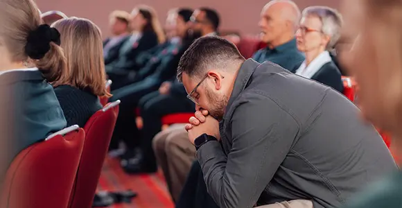 Man hunched over in prayer in the Assembly hall surrounded by others looking ahead.