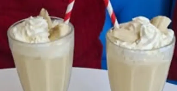 Two banana milkshakes topped with whipped cream and banana slices, served in tall glasses on a café table.