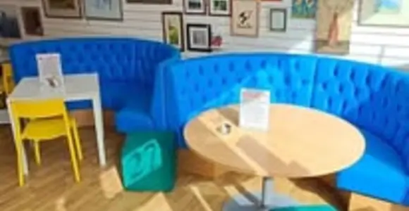 New seating area in a community café featuring blue upholstered booth seating, round wooden tables and colourful stools, with framed artwork displayed on the walls.
