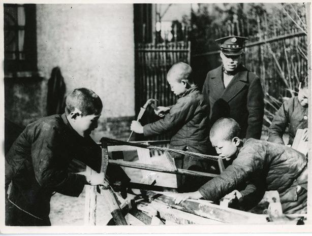 Children with cropped hair saw logs, overseen by man in uniform