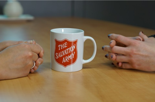 A mug with a Salvation Army logo in between two people's closed hands on a table