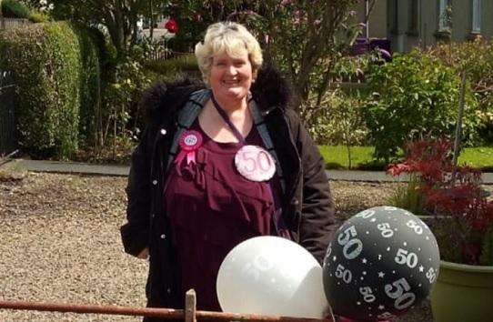 A photo of a blonde lady smiling wearing 50th birthday badges and holding 50th birthday balloons.