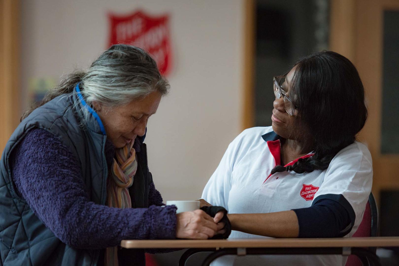 Two women are say together at a table. One woman, who is wear a Salvation Army polo shirt places her hand on the hands of the other women in reassurance.