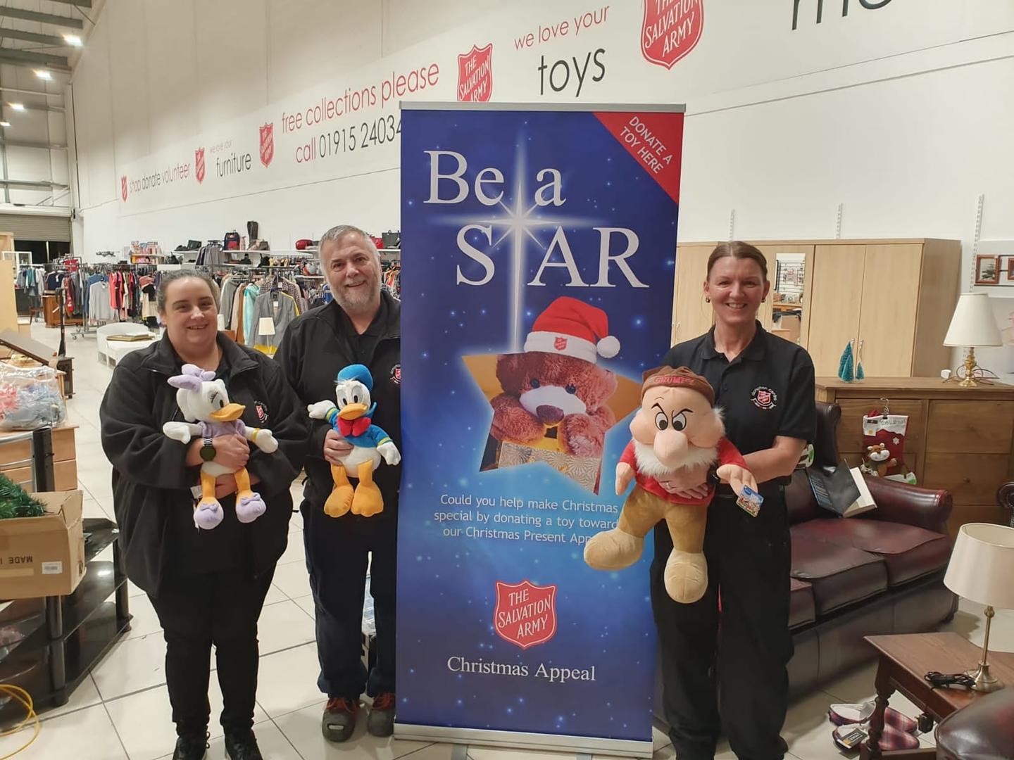 Three people in Salvation Army uniforms stand next to a 'Be a Star' campaign poster holding soft toys.