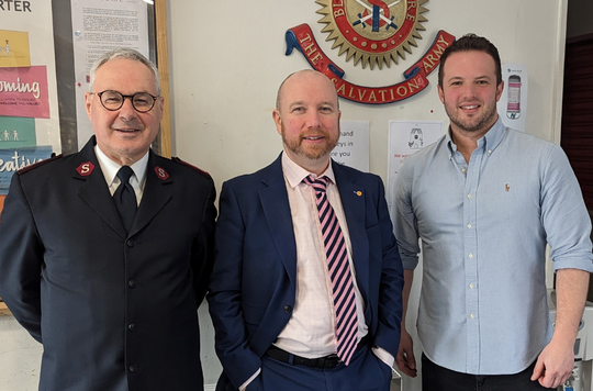 Major Roger Batt, Divisional Leader for Wales, Mabon ap Gwynfor, Member Senedd and Hugh Carter, Service Manager for Tŷ Gobaith standing in front of The Salvation Army emblem in Tŷ Gobaith Lifehouse.