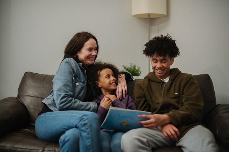 A woman sitting on the sofa with her children, she has her arms around her young daughter and they are both looking at and smiling at a teenage boy reading a kids book.