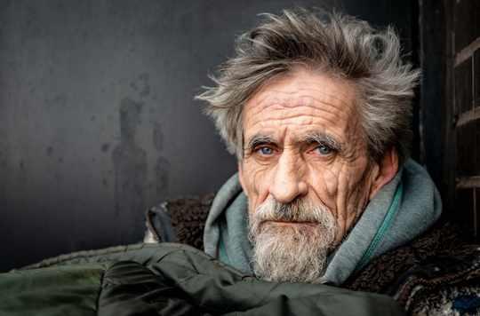 a close up image of the face of an older man sleeping rough on the streets, he has a long grey beard and is trying to keep warm in his sleeping bag.