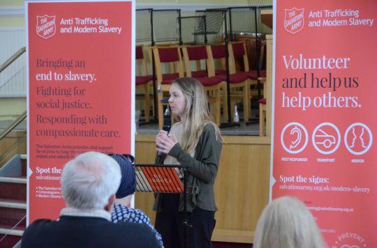 Abi Pitt, Mentoring Co-ordinator speaking to an audience at a modern slavery roadshow in Nottingham. Abi has long blonde hair and is holding a microphone while standing at a lectern.