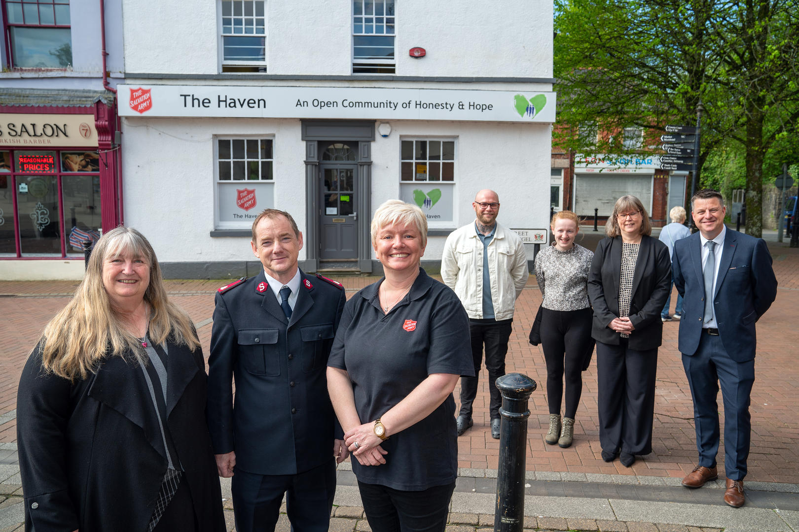 Welsh Minister Julie James, Salvation Army Officer Major Neil Duquemin and and Project Development Manager Christina Davies stand in a line. Four other individuals stand in a line nearby. Behind everyone is Neath Salvation Army's drop-in centre, The Haven