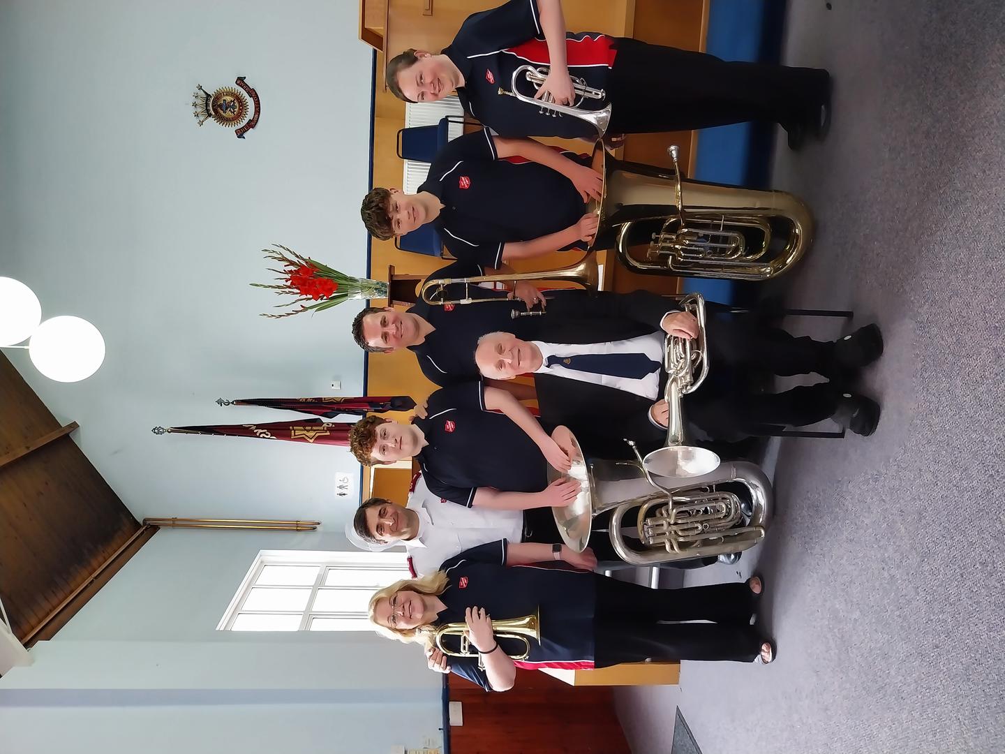A group of six brass players wearing Salvation Army polo shirts, with a corps officer in the middle standing in front of another brass player sitting down. 