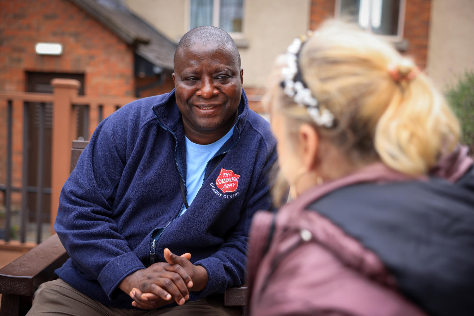 A male wearing a Salvation Army fleece with Granby Centre embroidered on it, talks with woman wearing purple coat