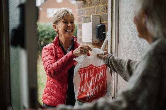 A Salvation Army volunteer wearing a red puffer jacket delivers a care package to an older woman