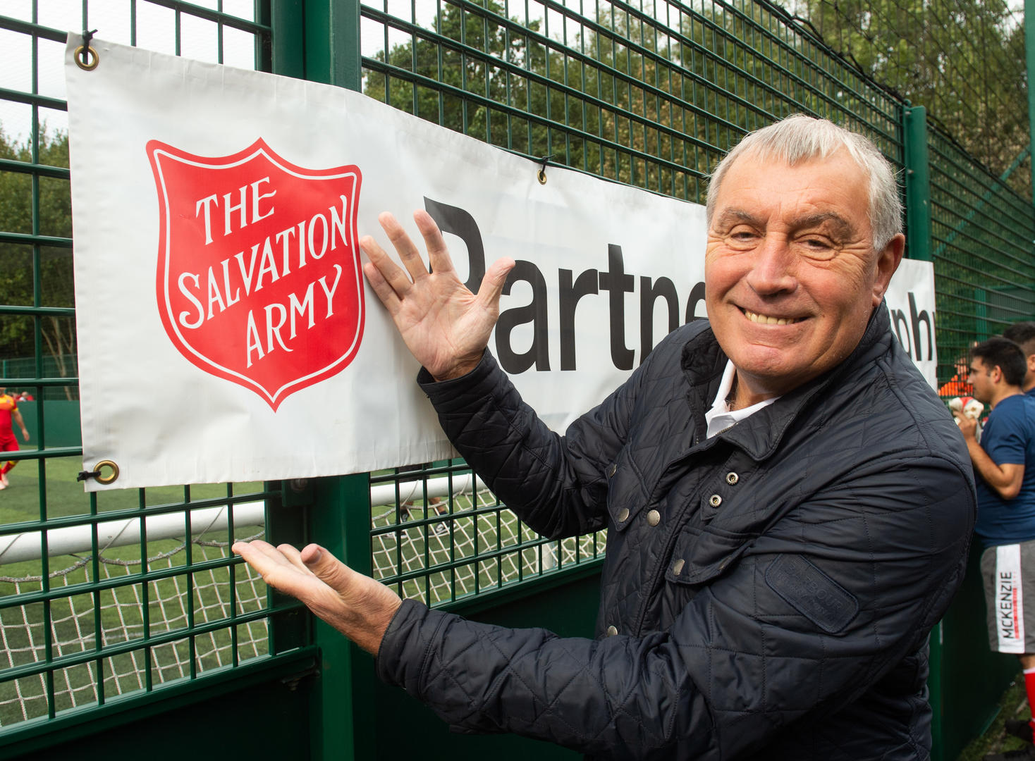 Peter Shilton standing next to the Partnership Trophy banner and pointing to the Salvation Army red shield.