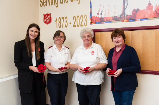 Two volunteers and two Salvation Army Officers are stood in a row presenting knitted red poppies