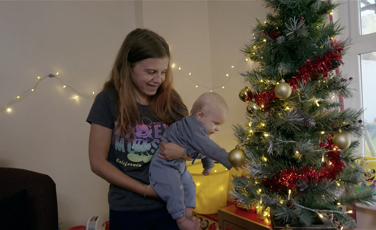 Connie is smiling as she holds her youngest child, they are both decorating a Christmas tree. There are fairy lights in the background.