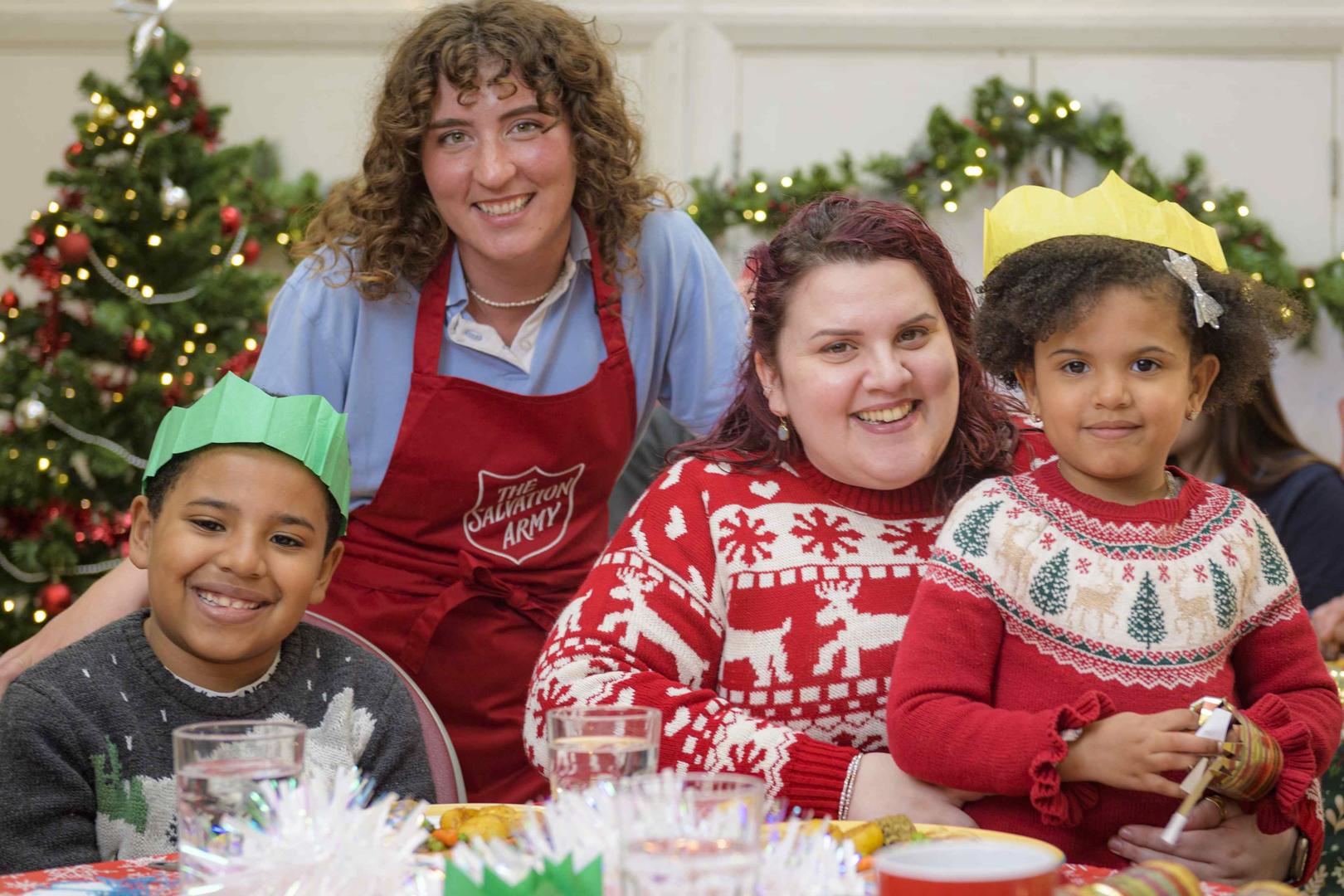 A mum and her two children a boy and a girl, wearing christmas jumpers, are enjoying a christmas lunch at a Salvation Army church. A volunteer wearing a red Salvation Army apron is standing with them and they are all looking at the camera and smiling. 