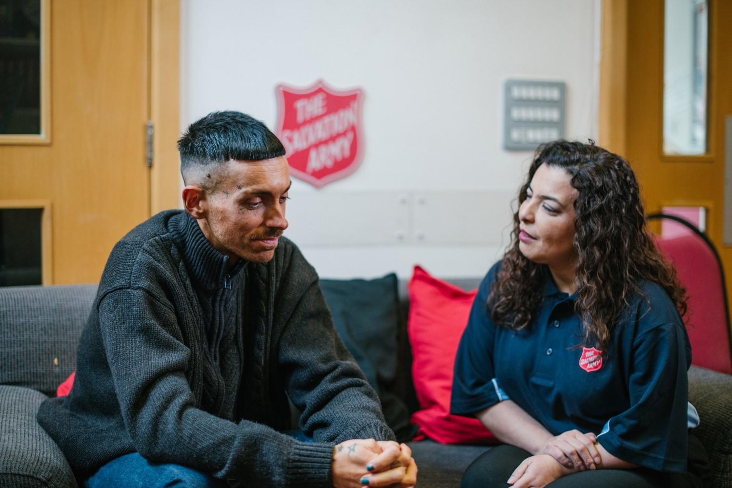 A Salvation Army volunteer/staff member talking with Max on a grey sofa inside a Salvation Army building.