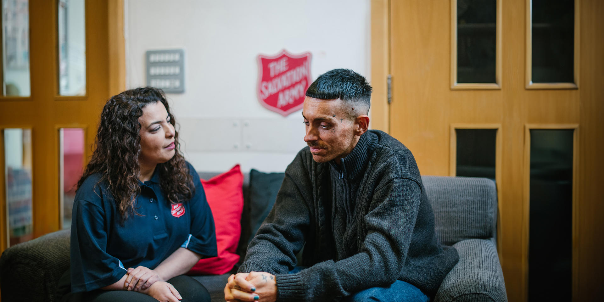 A Salvation Army volunteer/staff member talking with Max on a grey sofa inside a Salvation Army building.