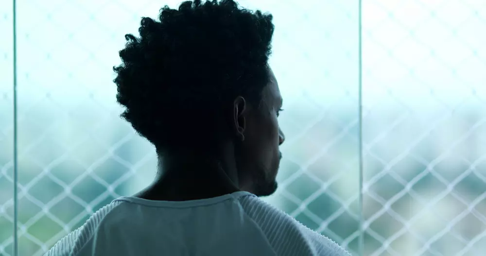 A bright image of a young black man looking out of a window, you cant see his face, he is wearing a white t-shirt. 