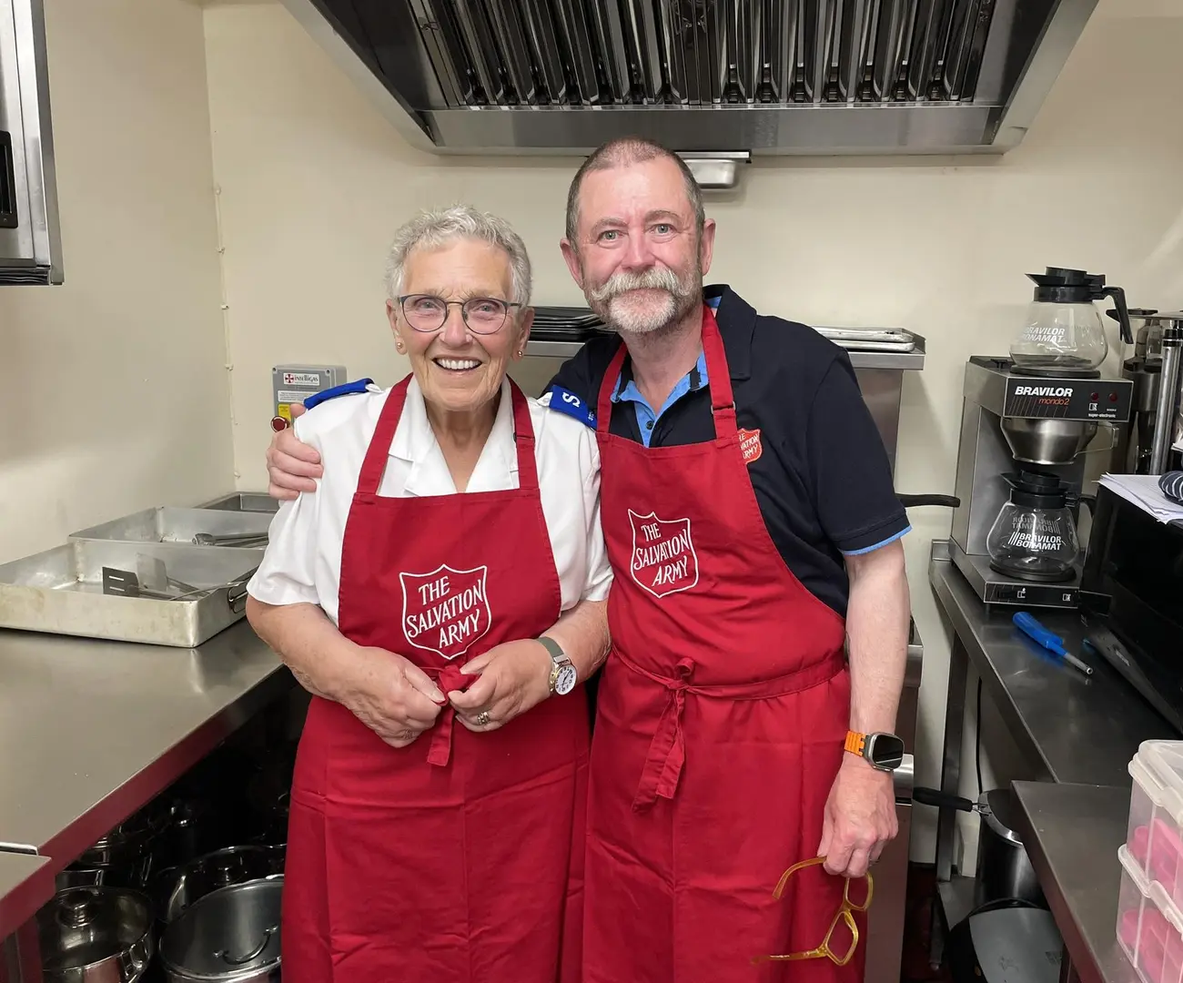 Two smiling Salvation Army volunteers wearing aprons with a red shield stand together in a kitchen, with one person’s arm gently around the other.