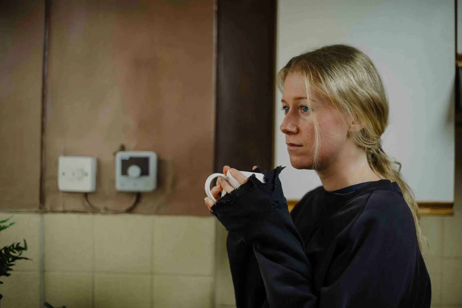 A person with long blonde hair and a dark, frayed-sleeve sweater holds a white mug near their face. The background includes a beige wall with mounted electrical devices and green plants.