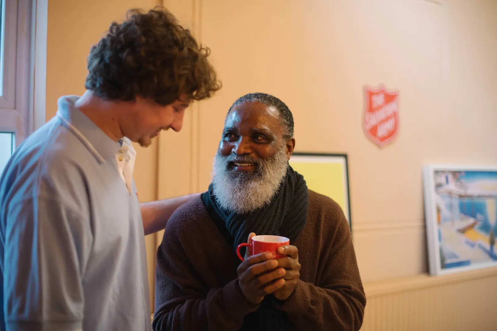 Man smiling towards the camera, wearing a red jumper and holding a cup of tea.