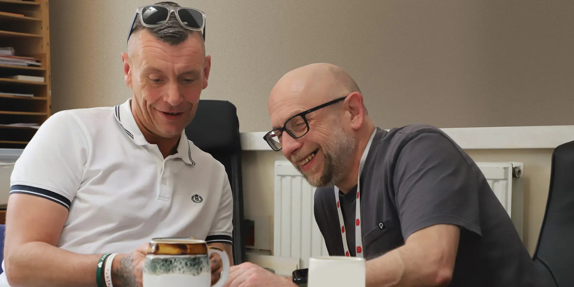 Two men sat at a table, one is showing the other something of interest. Both are joyful and smiling.