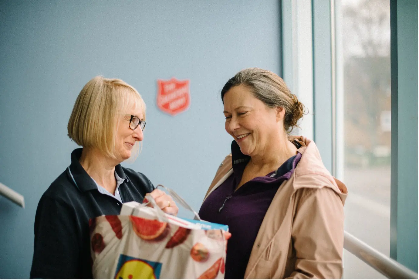 Two women smiling at eachother, a salvation employee handing a bag of shopping to another woman. A faint Salvation Army logo can be seen on the wall behind.