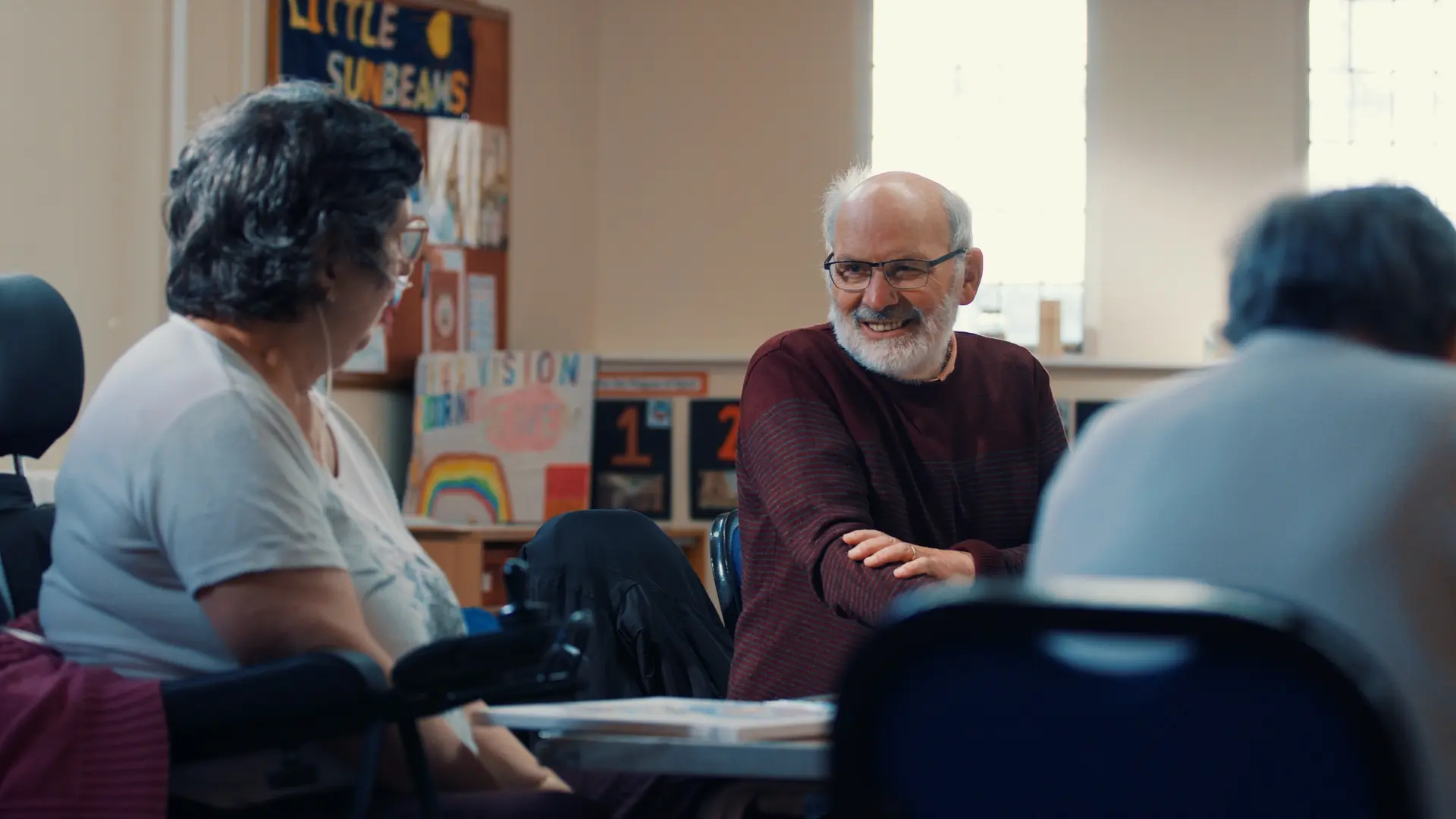 Three people are seated in a well-lit room having a conversation. On the left, one person is in a wheelchair. In the centre, another person with sits with arms crossed. A third person is partially visible on the right.