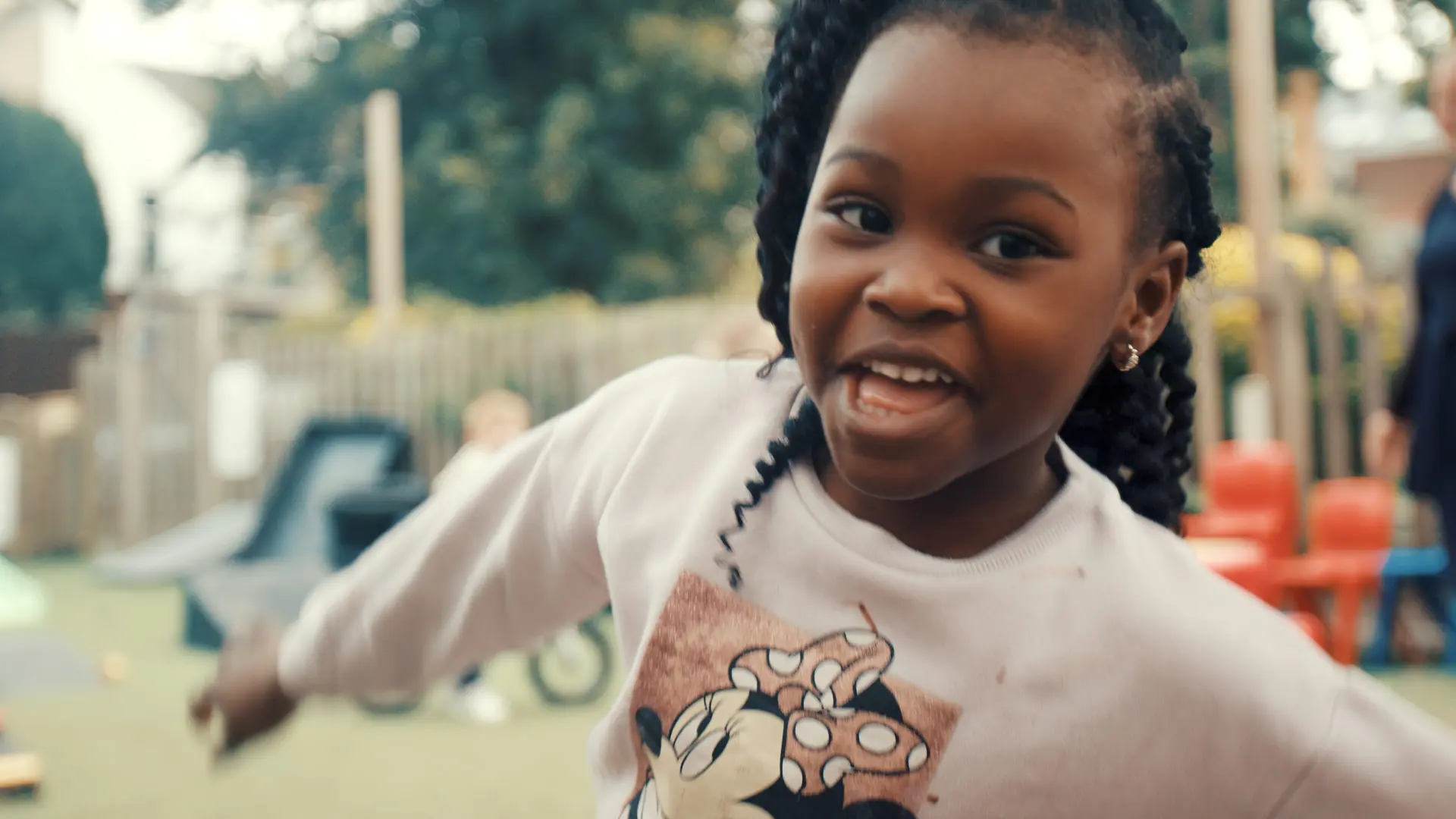 A child wearing a white sweatshirt with a Minnie Mouse graphic plays outdoors in a playground area with various equipment and other children.