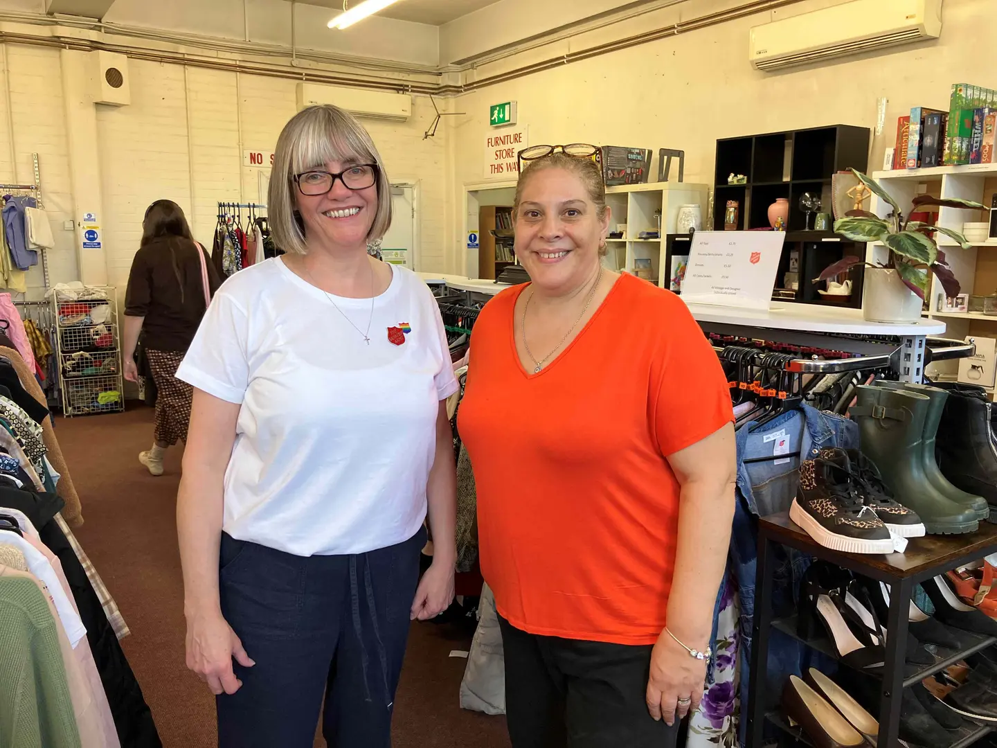 Church leader, Julie Pell and charity shop manager Oya standing in 'Sally in the Stow' charity shop. They are both smiling. Julie is wearing glasses and a white t-shirt and Oya has a bright orange t-shirt