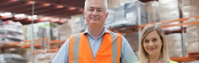 A man and a woman wearing high visibility jackets, standing infront of a warehouse.
