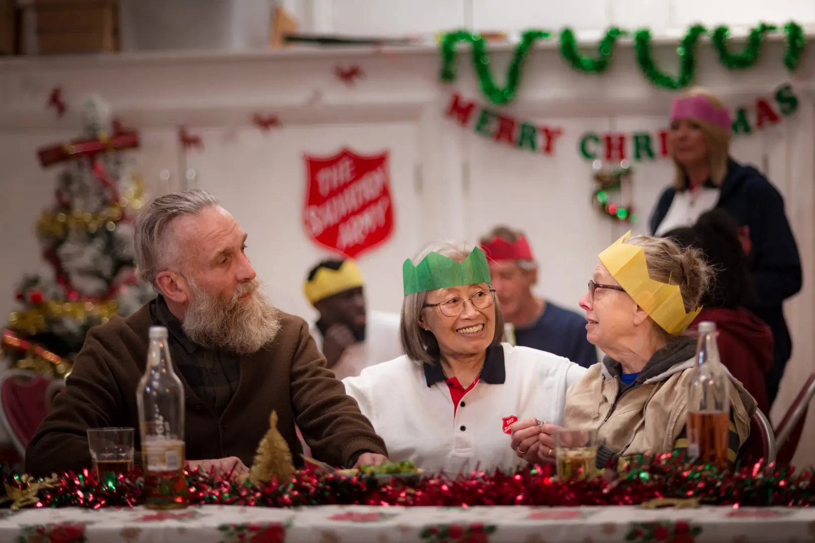 A man speaking to two women, each wearing christmas hats. The Salvation Army logo is visible in the background next to christmas decorations. All people look happy and festive.