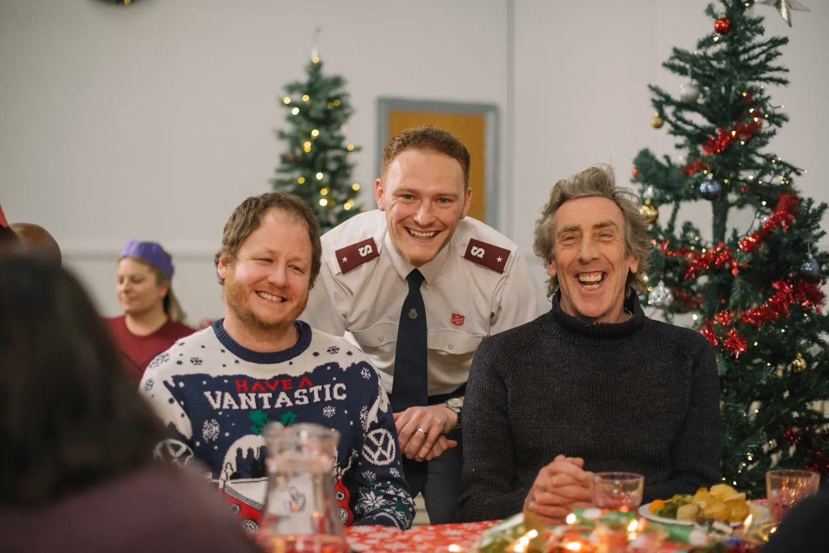 Two men alongside a Salvation Army member, enjoying a festive christmas lunch. Everyone looks happy.