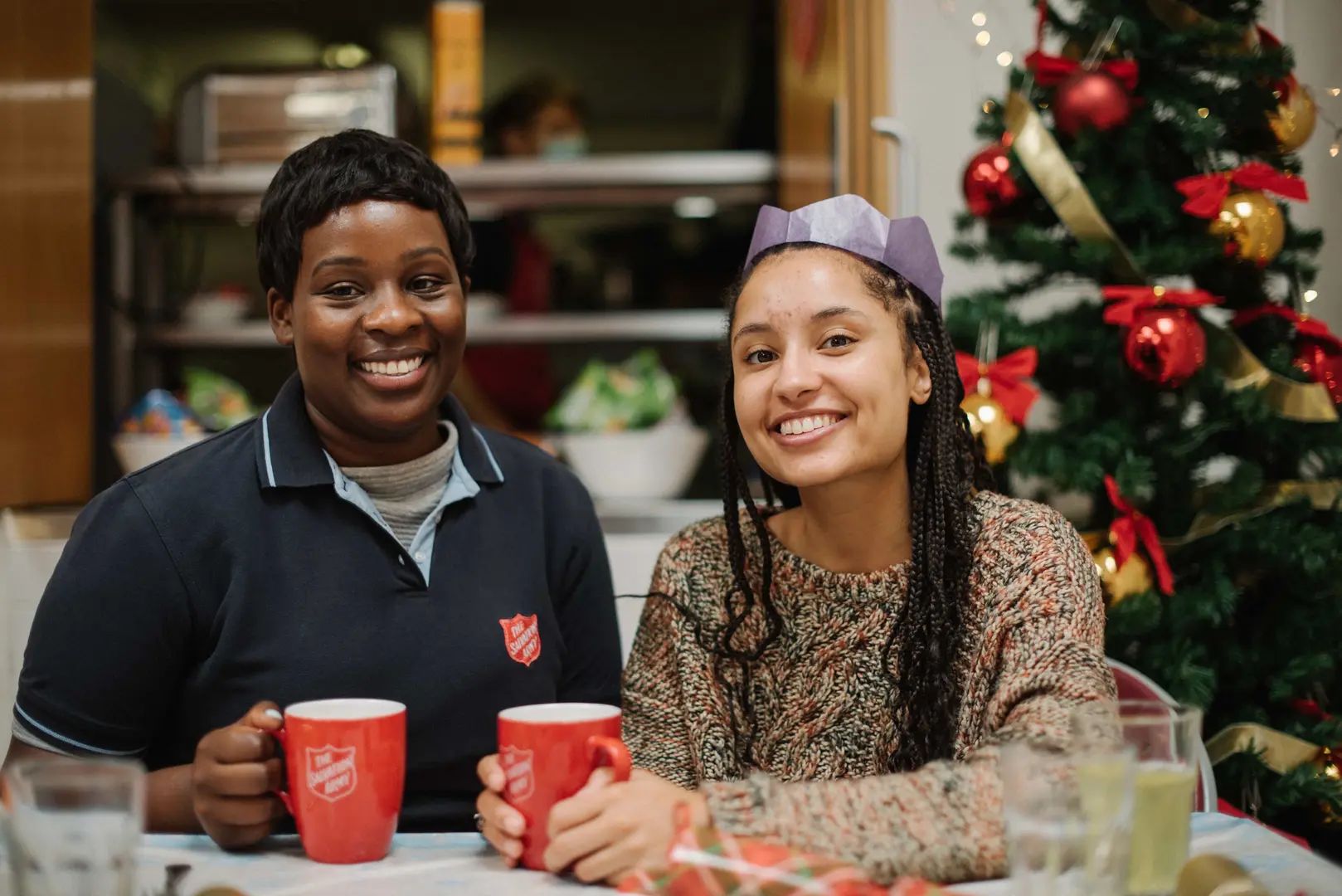 Two women enjoying a cup of tea, at a festive gathering with a christmas tree in the background. Both women look very happy and festive.