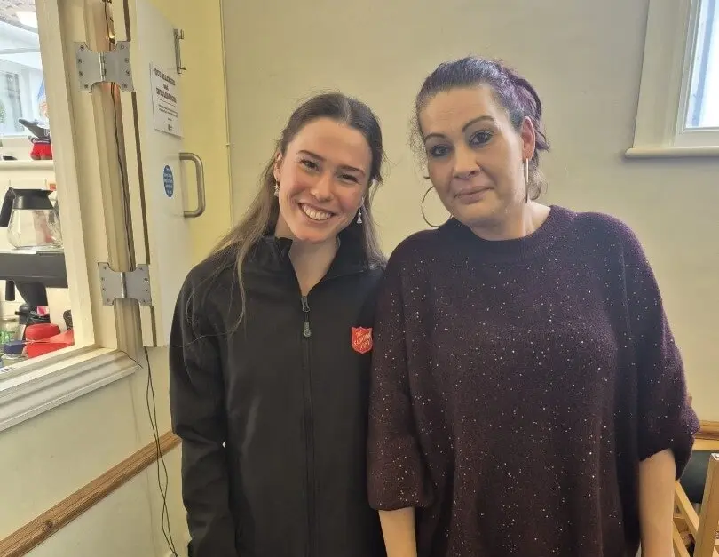A lady wearing a purple sparkly jumper is pictured standing next to a Salvation Army officer. Both people are smiling for the camera. 