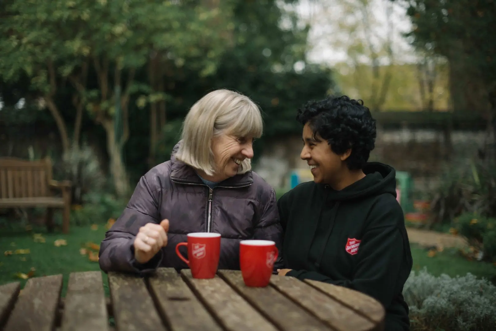 Two women sitting at an outdoor table, looking happy and deep in conversation. One is a salvation army employee, they are both drinking a hot drink.