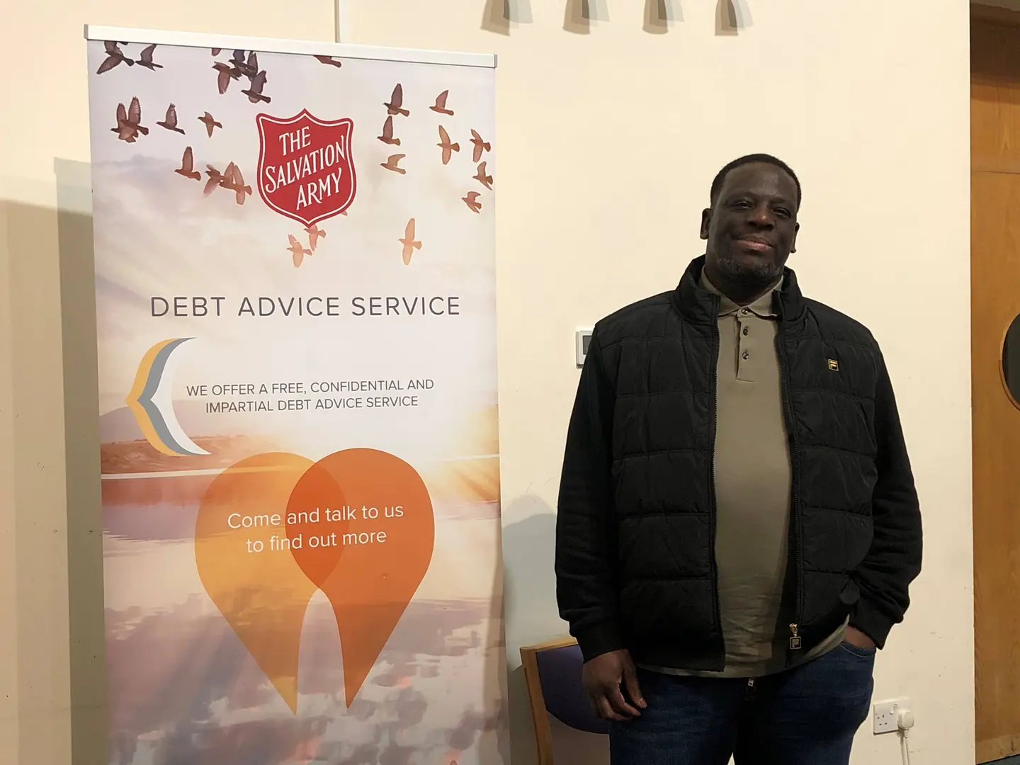 Ivan stands next to a sign advertising the debt advice service, he is relaxed with his hands in his pockets and smiles at the camera. 