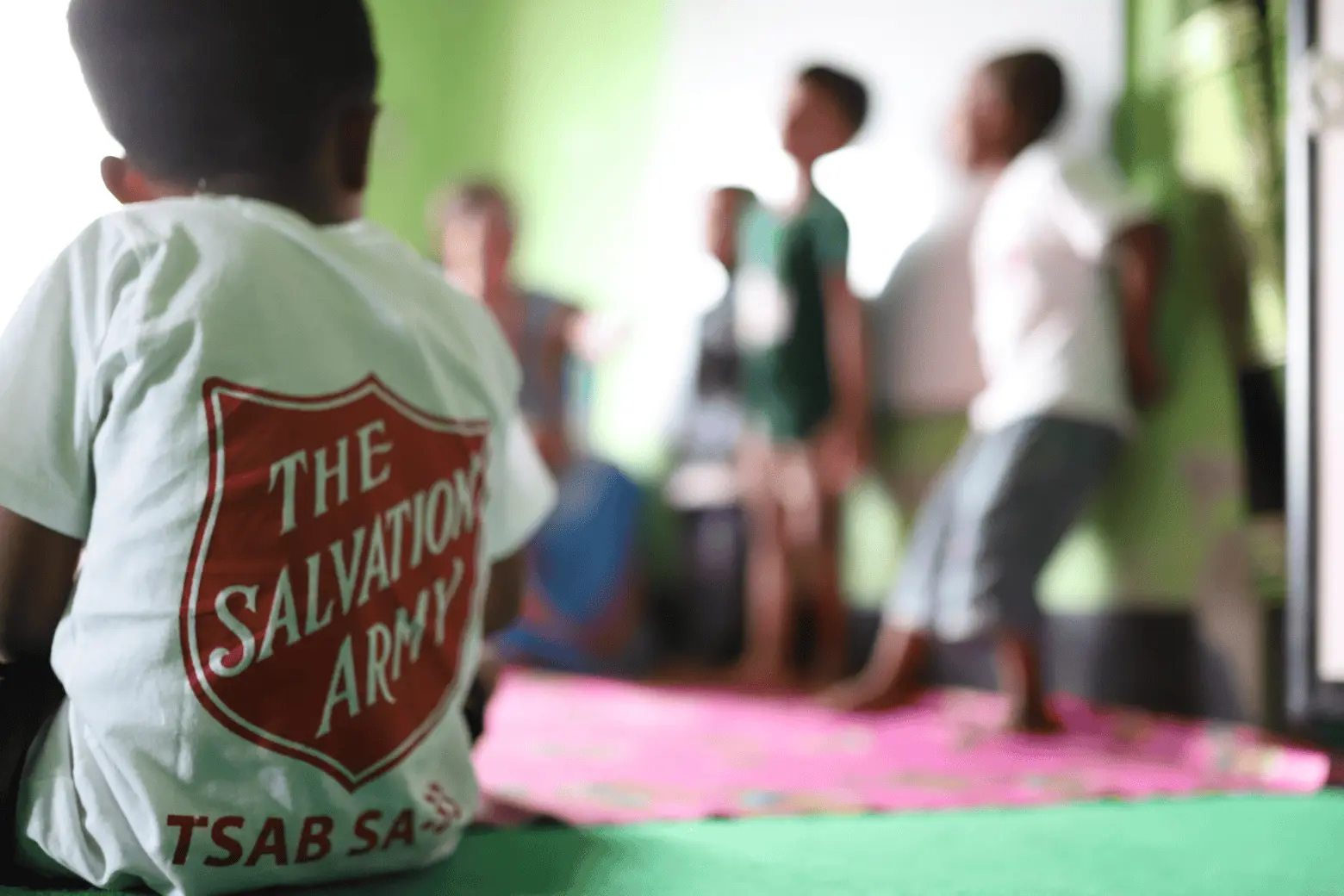 A child wearing a Salvation Army T‑shirt sits on the floor watching a group of children standing at the front of a room.