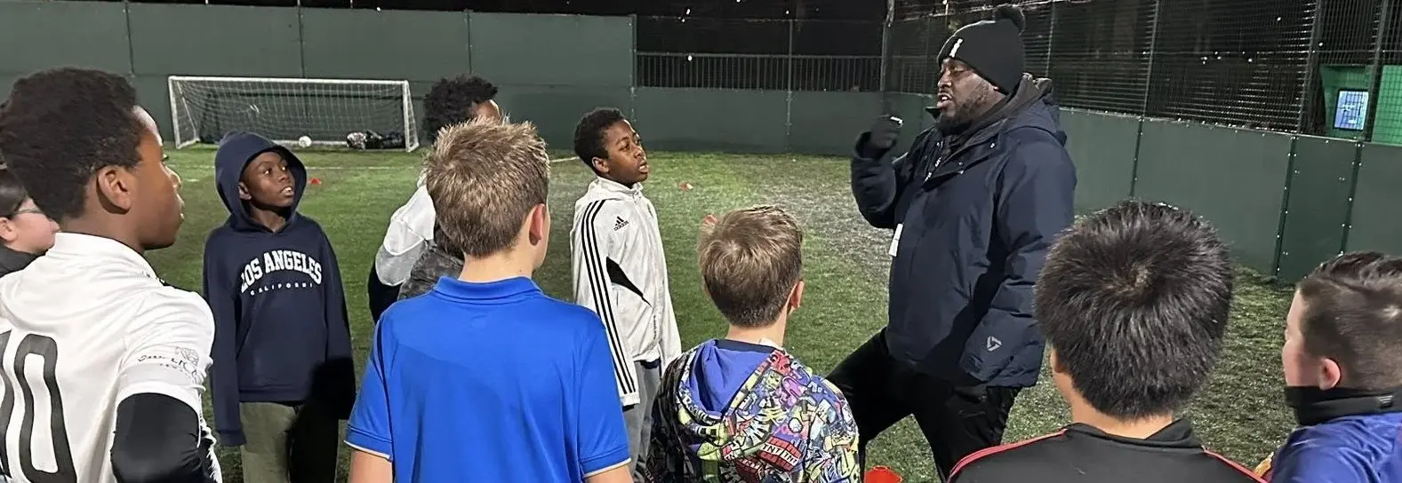A coach stands on an outdoor football pitch at night, speaking to a group of children gathered in a semicircle around him. The players wear sports clothing, and a goalpost is visible in the background under the pitch floodlights.