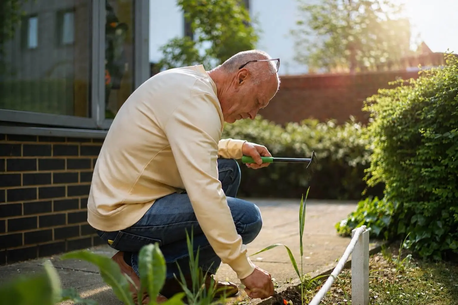 A man wearing a white jumper is kneeling down to put plants into a garden.