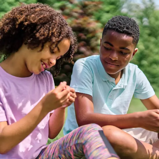 Two children sitting on grass in a sunny outdoor setting, closely examining a small object together, surrounded by green plants.