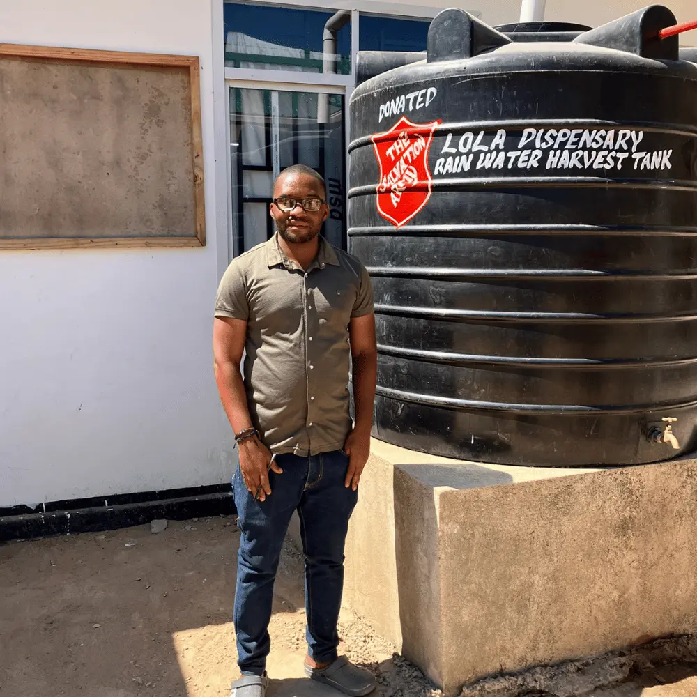 A man (Dr Leonard Mfugale) stands beside a large rainwater harvesting tank at Lola Dispensary in Tanzania