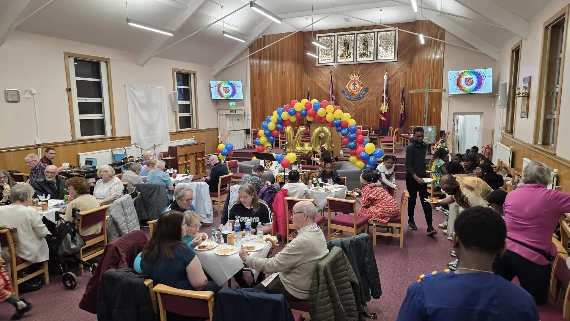 This image shows the inside of the main hall set out cafe style tand is full of church people celebrating with food and refreshments on the 140th anniversary. It is a very colourful image.